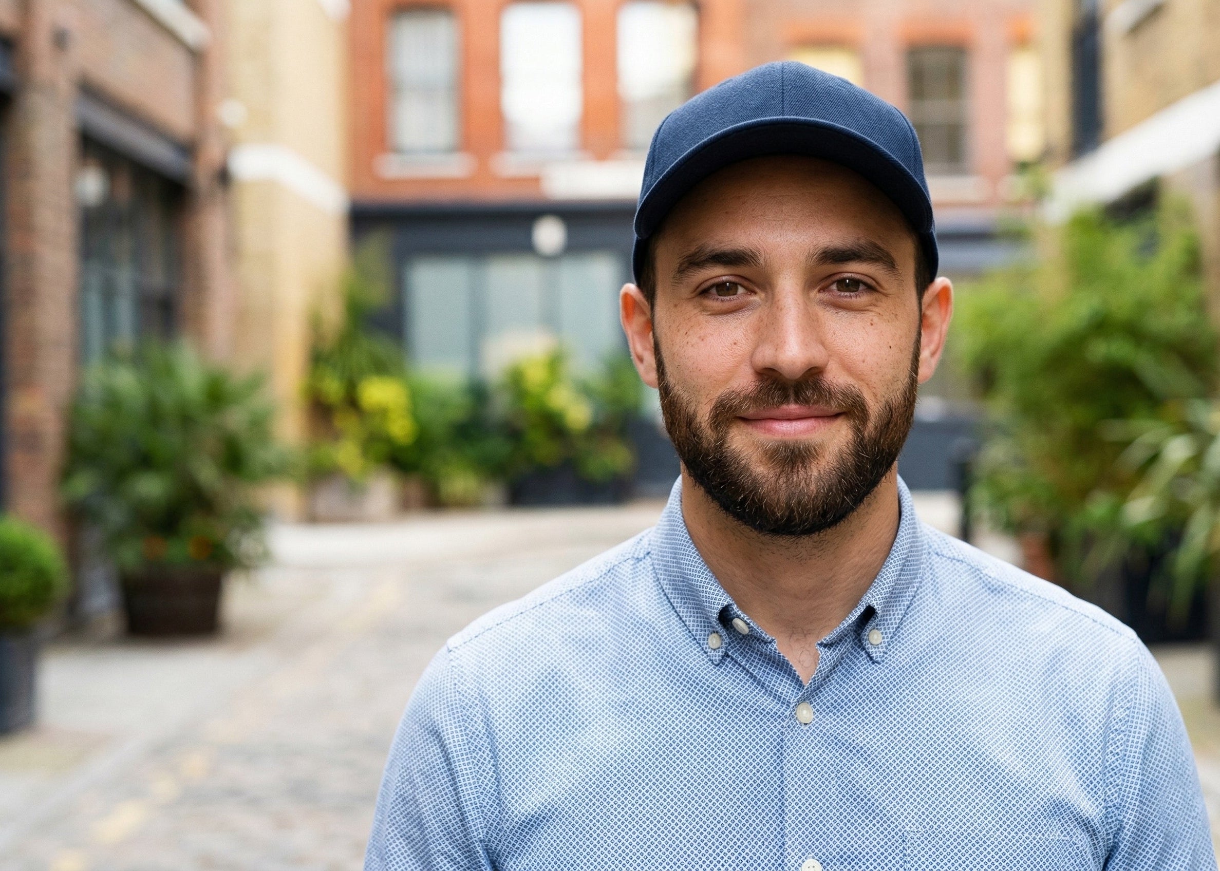 A smiling young man in a blue baseball cap standing in an urban alleyway. As a dedicated user of iFORWAY portable power solutions, he represents urban creators and travelers who need to keep their cameras and devices powered throughout the day.