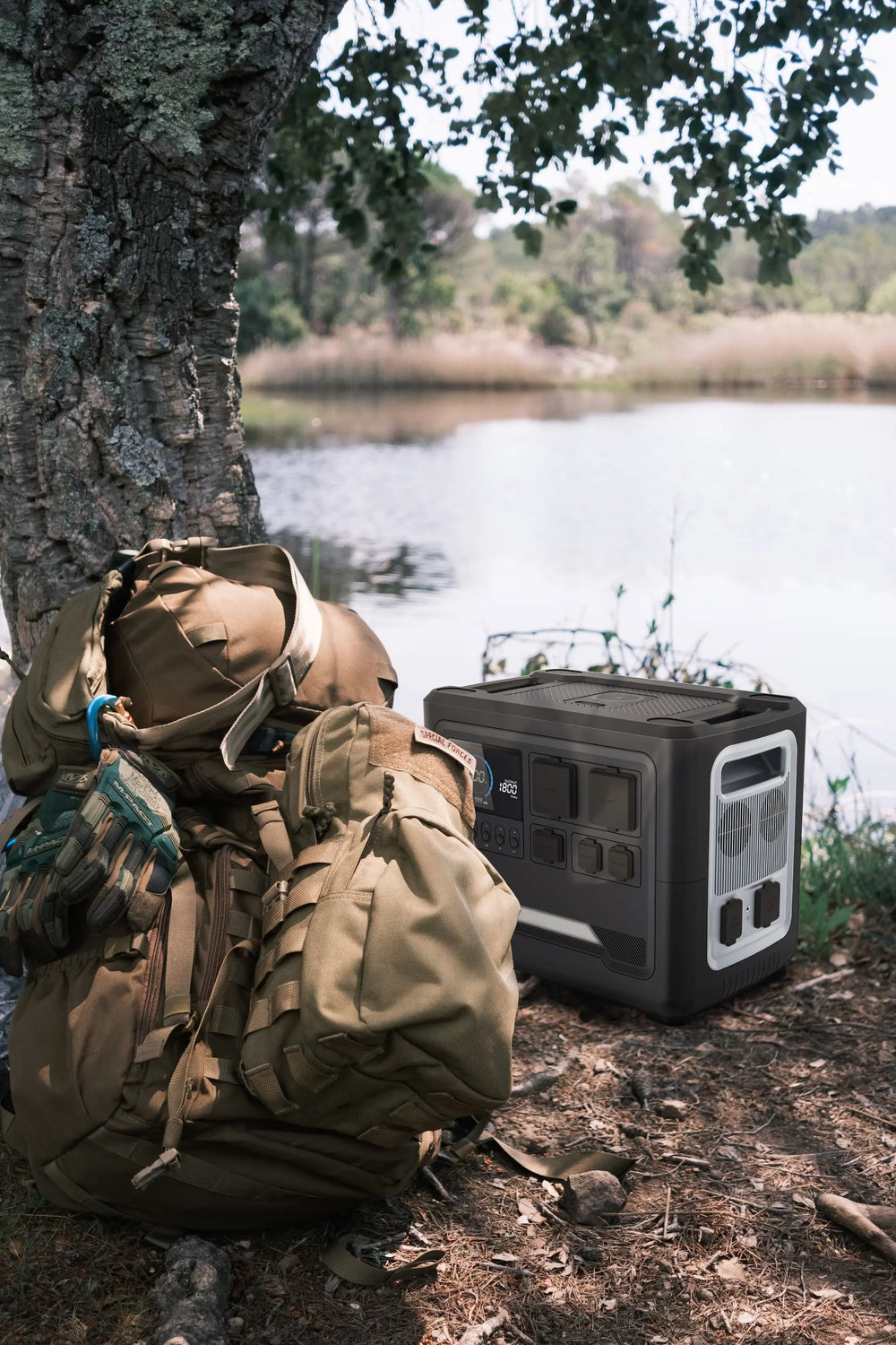 An iFORWAY high-capacity portable power station next to a tactical backpack by a peaceful lake. This scene highlights the product's reliability for wilderness survival, hunting, or fishing trips, serving as a robust off-grid power hub for camping gear and essential electronics.