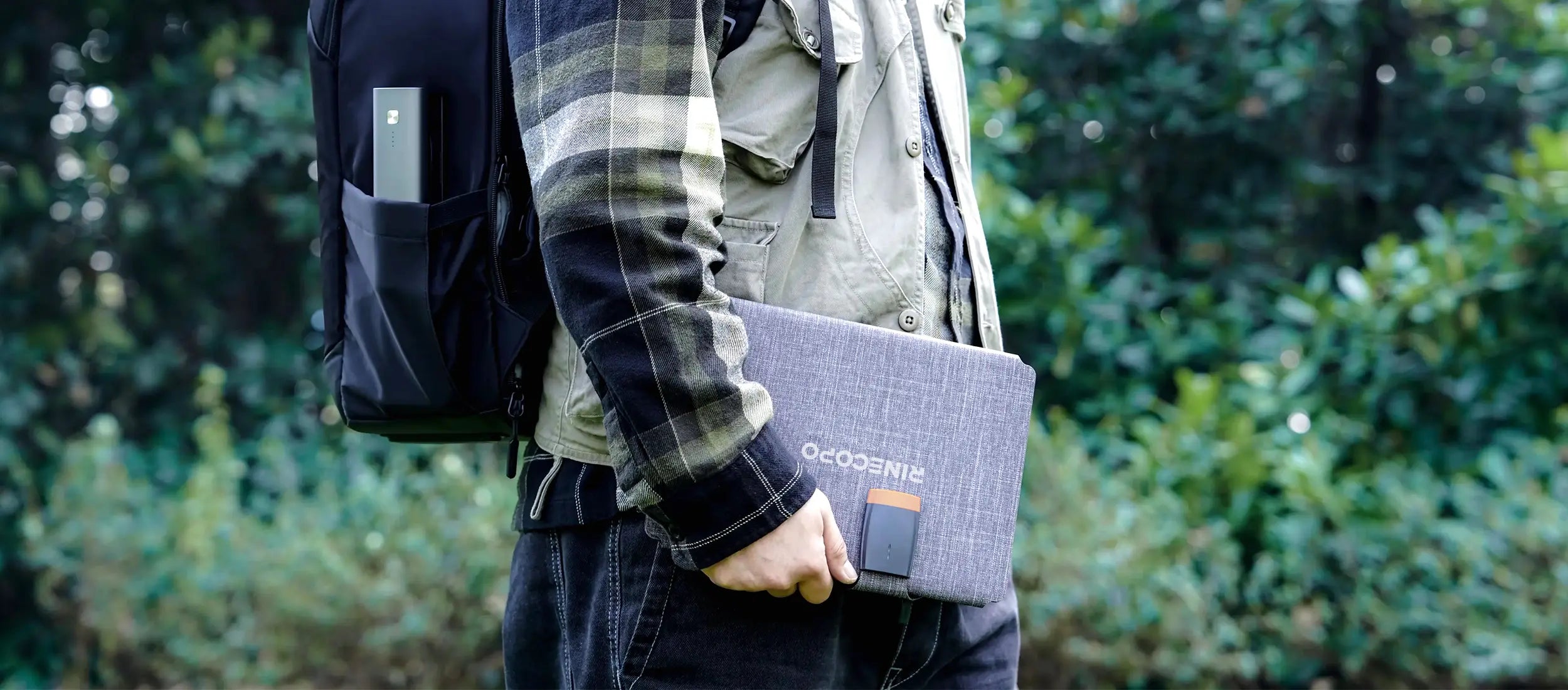 A hiker walking through the woods carrying a folded RINECOPO portable solar charger. Built tough to be waterproof, heat-resistant, and corrosion-proof, this lightweight solar panel features fast USB-C charging, making it the perfect power-on-the-go solution for camping, trekking, and outdoor adventures.